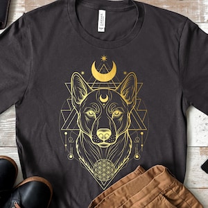 以下が含まれることがあります： 犬の頭、三日月、星の幾何学模様が金色のダークグレーのTシャツ。犬の顔は、三角形と円のネットワークの中心にあります。柔らかい素材で作られたシャツです。