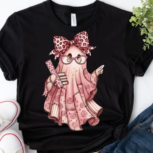 Puede incluir: Camiseta negra con un fantasma rosa que lleva gafas y un lazo con diseño de estampado de leopardo. El fantasma sostiene libros y una regla. El fantasma lleva un atuendo rosa patchwork con patrones de rosas.