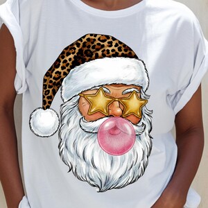 Puede incluir: Camiseta blanca con un gráfico de Papá Noel. Papá Noel lleva gafas de sol en forma de estrella y un gorro con estampado de leopardo. Una gran burbuja de chicle rosa está en su boca. La barba de Papá Noel es blanca.