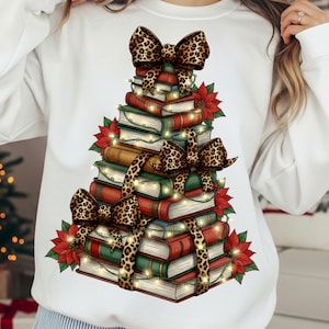 May include: White sweatshirt featuring a Christmas tree design made of books. The books are stacked and adorned with leopard print bows, red poinsettias, and string lights. A festive and cozy design for the holiday season.