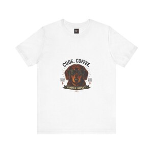 May include: A white short-sleeve t-shirt featuring a graphic of a brown dachshund wearing black sunglasses. The text reads CODE. COFFEE. FOCUS MODE. ZERO BUGS. COMPILE. REPEAT. with a small code bracket symbol at the bottom of the design.