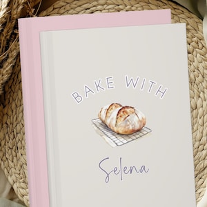 May include: A light gray notebook with a pink border features the text "Bake With Selena" above a watercolor illustration of a loaf of bread on a cooling rack. The notebook is resting on a woven mat.