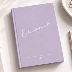 Puede incluir: Un diario de color lavanda con el nombre "Eleanor" en escritura cursiva. Un pequeño corazón está debajo de una línea horizontal en la parte inferior. También se ven un bolígrafo dorado, un plato pequeño con joyas y un par de gafas.