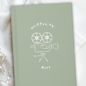 May include: Sage green notebook with a white sketch of a vintage film camera and the words "written by:" above it. The name "Alex" is written below the camera. The notebook is on a white surface with white flowers.