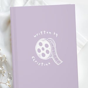 May include: A lavender journal with the words "written by Christina" and a film reel illustration in white. The journal is closed and resting on a white surface with floral accents.