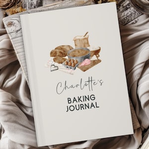 Puede incluir: Un diario de cocina crema con el título "Charlotte's Baking Journal" en escritura negra. La portada presenta una ilustración de acuarela de ingredientes para hornear, incluyendo pan, huevos y harina.