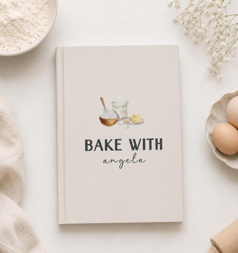 Puede incluir: Un libro de cocina beige con el texto "BAKE WITH angela" en negro. La portada presenta ilustraciones de acuarela de ingredientes para hornear: un cuenco con un batidor, una bolsa de harina, mantequilla y huevos. El libro est&aacute; sobre una superficie blanca.