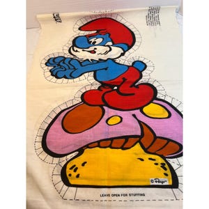 May include: A white fabric panel featuring a printed Smurf character wearing a red hat and red pants, standing on a large pink and yellow mushroom. The design includes dashed cutting lines, sewing instructions, and the text LEAVE OPEN FOR STUFFING.