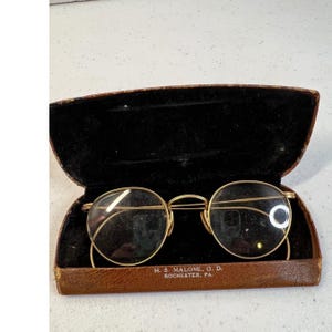 May include: Gold-tone round wire-rimmed glasses with thin temples resting in a brown leather-textured case with black velvet lining. The case features the printed text H. S. MALONE, O. D. ROCHESTER, PA. on the interior bottom edge.