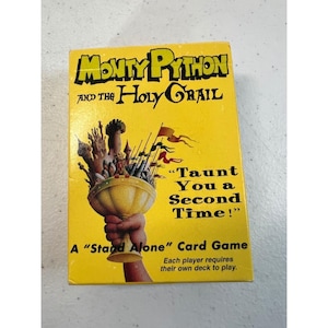 May include: A yellow rectangular card game box titled Monty Python and the Holy Grail. The cover features a hand holding a golden chalice filled with castle towers and flags, with the text Taunt You a Second Time! and A Stand Alone Card Game.