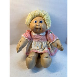 Peut inclure: Une poupée en tissu avec une peau claire, des yeux bleus et des cheveux en laine blonde bouclée. La poupée porte une robe à carreaux vichy rose et blanc avec un col blanc, une bordure en dentelle et une petite étiquette verte sur la manche gauche.