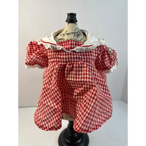 May include: A red and white gingham dress featuring short puff sleeves, a wide white collar with delicate eyelet lace trim, and a gathered skirt. The garment is displayed on a mannequin torso, showcasing the classic checkered pattern.