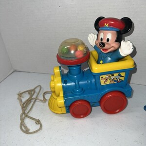 May include: A plastic pull-toy train featuring a Mickey Mouse figure in a blue uniform and red cap. The train is blue and yellow with red wheels, a clear dome containing colorful small balls, and a sticker depicting Pluto and Donald Duck.