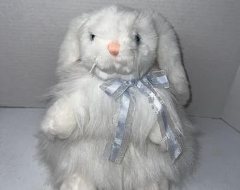 May include: A plush white rabbit toy with long, soft fur, featuring a small pink nose, blue eyes, and white whiskers. The rabbit is adorned with a decorative, translucent light blue ribbon tied in a bow around its neck, resting against its chest.