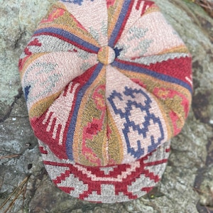 May include: A multi-panelled newsboy cap featuring a woven tapestry-style fabric with a geometric pattern in shades of red, beige, blue, and gold. The cap has a central fabric-covered button on the crown and a structured brim with a matching pattern.