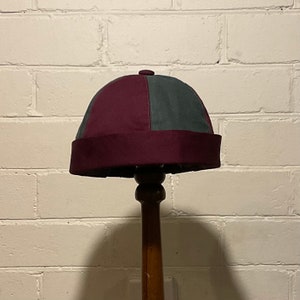 Peut inclure: Un chapeau bicolore avec un bord marron et un dessus marron et vert. Le chapeau est présenté sur un support en bois contre un mur de briques blanches. Le chapeau semble être fait de tissu.
