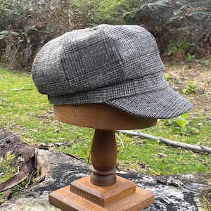 May include: A newsboy cap featuring a grey and black plaid pattern with a structured crown and a short, stiff brim. The cap is displayed on a wooden stand, showcasing the textured fabric weave and the classic multi-paneled construction of the hat.