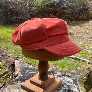 May include: A solid burnt orange newsboy cap featuring a soft, suede-like texture, a rounded crown with multiple panels, and a short, stiff brim with horizontal stitching detail. The cap is displayed on a wooden stand to show its shape.