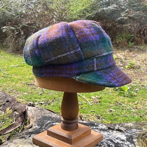 May include: A baker boy cap featuring a multi-coloured plaid pattern in shades of purple, green, and brown. The cap has a rounded, panelled crown and a short, stiff brim, constructed from a textured wool-like fabric with a visible weave throughout.
