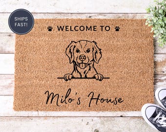 May include: A rectangular coir doormat with a dog illustration and the text "WELCOME TO" above the dog and "Milo's House" below. The mat is brown with black text and a black dog outline.