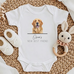 May include: A white short-sleeved baby bodysuit featuring a graphic of a golden retriever dog above a horizontal black line. Below the line, the text reads "Oscar's NEW BEST FRIEND" in black lettering. The garment has a round neckline and snaps.