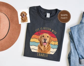 May include: Dark gray t-shirt featuring a golden retriever dog with the text "LIFE IS BETTER WITH CHARLIE" in a retro sunset design. A small photo of the dog is also present. A brown hat and blue jeans are in the background.