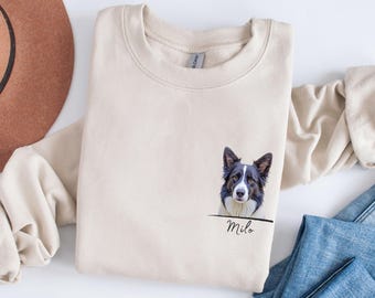 May include: A cream-colored sweatshirt featuring a detailed portrait of a Border Collie dog. The dog's image is on the front, with the name "Milo" printed below. The sweatshirt is folded, with a brown hat and blue jeans visible.