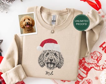 May include: A beige sweatshirt featuring a graphic of a dog wearing a Santa hat, with the name "Milo" below. A framed photo of a dog is on the left. The sweatshirt has a crew neck and long sleeves. A green circle with white text says "UNLIMITED PROOFS!"