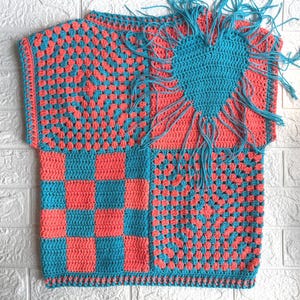 Puede incluir: Una prenda superior de ganchillo sin mangas con un diseño de patchwork en hilo color coral y turquesa. La pieza incluye un patrón de granny square, un bloque a cuadros y un gran motivo de corazón en la parte superior derecha con flecos.