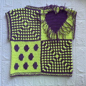 Könnte beinhalten: Ein ärmelloses Häkeloberteil mit einem Vier-Panel-Design in Limettengrün und Dunkelviolett. Das Kleidungsstück besteht aus zwei Granny-Square-Feldern, einem Feld mit violetten Punkten und einem Feld mit einem großen violetten Herz mit Fransen.