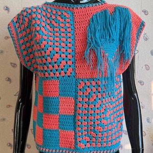 Peut inclure: Un haut sans manches en crochet présentant un design color-block en corail et turquoise. Le vêtement combine des motifs de carrés granny, des sections en brides pleines et un quadrillage, agrémenté d'une frange décorative turquoise.