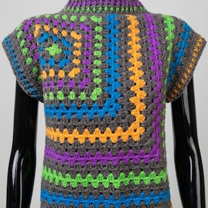 Op de afbeelding: Een gehaakte top met korte mouwen en een vierkant patroon in antracietgrijs, geaccentueerd met levendige strepen in neongroen, felblauw, paars en oranje. Het kledingstuk heeft een hoge halslijn en een textuur die met de hand is gemaakt.