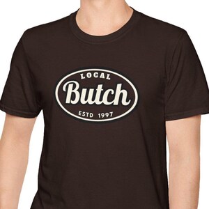 Op de afbeelding: Een donkerbruin T-shirt met korte mouwen en een centraal ovaal grafisch ontwerp met een zwarte achtergrond en een witte rand. De tekst in het ovaal luidt "LOCAL" bovenaan, "Butch" in een groot cursief lettertype in het midden en "ESTD 1997" onderaan.