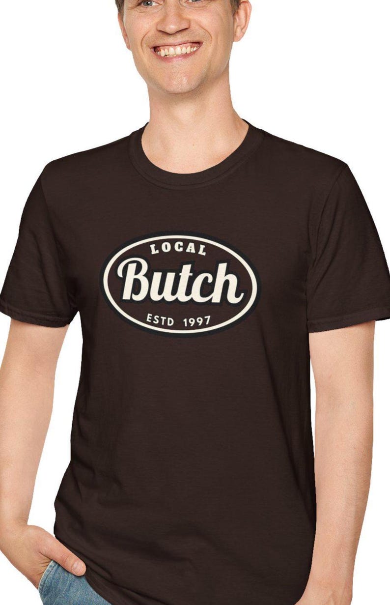 Op de afbeelding: Een donkerbruin T-shirt met korte mouwen en een centraal ovaal grafisch ontwerp met een zwarte achtergrond en een witte rand. De tekst in het ovaal luidt "LOCAL" bovenaan, "Butch" in een groot cursief lettertype in het midden en "ESTD 1997" onderaan.