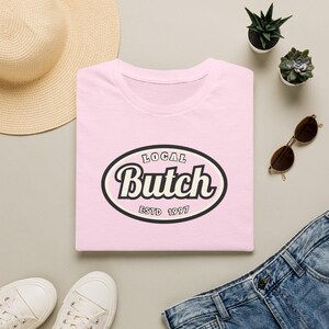 Op de afbeelding: Een lichtroze t-shirt met een centrale ovale afbeelding met een zwarte rand. De tekst binnenin leest "LOCAL" bovenaan, "Butch" in grote witte schrijfletters in het midden, en "ESTD 1997" onderaan, alles op een cr&egrave;mekleurige achtergrond.