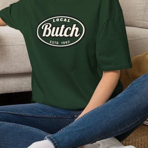 Op de afbeelding: Een donker bosgroen t-shirt met korte mouwen en een centrale ovale afbeelding met een zwarte rand. De tekst in het ovaal luidt "LOCAL" bovenaan, "Butch" in een groot wit cursief lettertype in het midden en "ESTD 1997" onderaan.