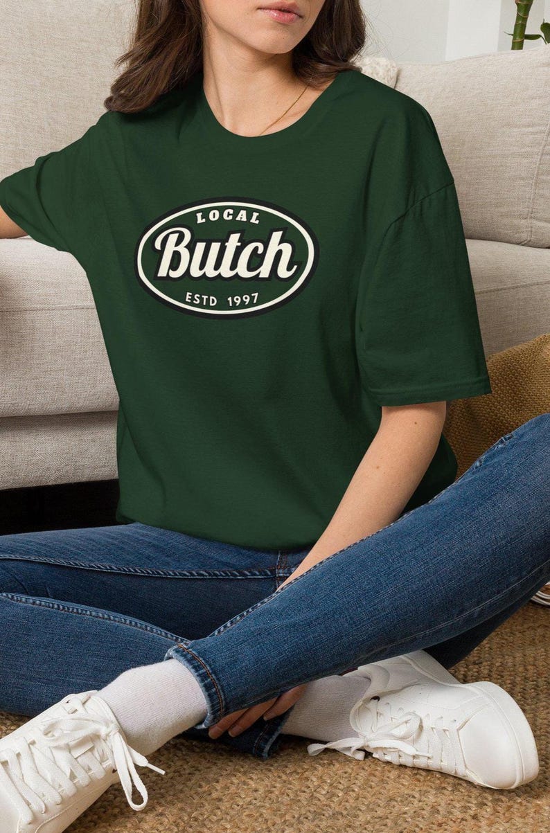 Op de afbeelding: Een donker bosgroen t-shirt met korte mouwen en een centrale ovale afbeelding met een zwarte rand. De tekst in het ovaal luidt "LOCAL" bovenaan, "Butch" in een groot wit cursief lettertype in het midden en "ESTD 1997" onderaan.