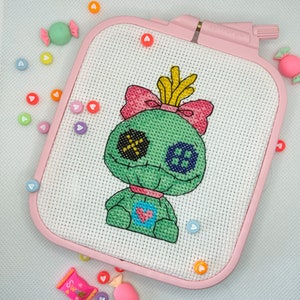 May include: A cross-stitch project featuring a green doll-like character with black, blue, and yellow details, set in a pink rectangular hoop. The design includes a pink bow and heart-shaped accents. Candy-shaped embellishments and heart-shaped beads surround the hoop.