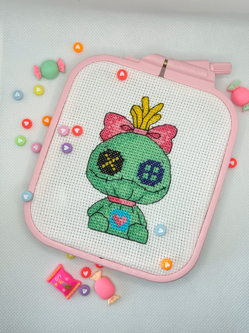 May include: A cross-stitch project featuring a green doll-like character with black, blue, and yellow details, set in a pink rectangular hoop. The design includes a pink bow and heart-shaped accents. Candy-shaped embellishments and heart-shaped beads surround the hoop.