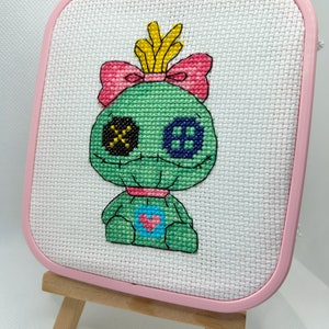 May include: A cross-stitch piece featuring a green creature with a pink bow, yellow crown, and heart detail. The artwork is displayed in a pink square hoop, mounted on a small wooden easel.