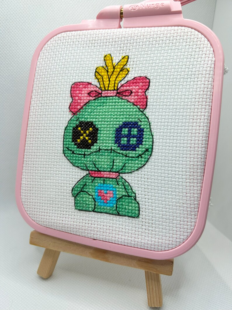 May include: A cross-stitch piece featuring a green creature with a pink bow, yellow crown, and heart detail. The artwork is displayed in a pink square hoop, mounted on a small wooden easel.