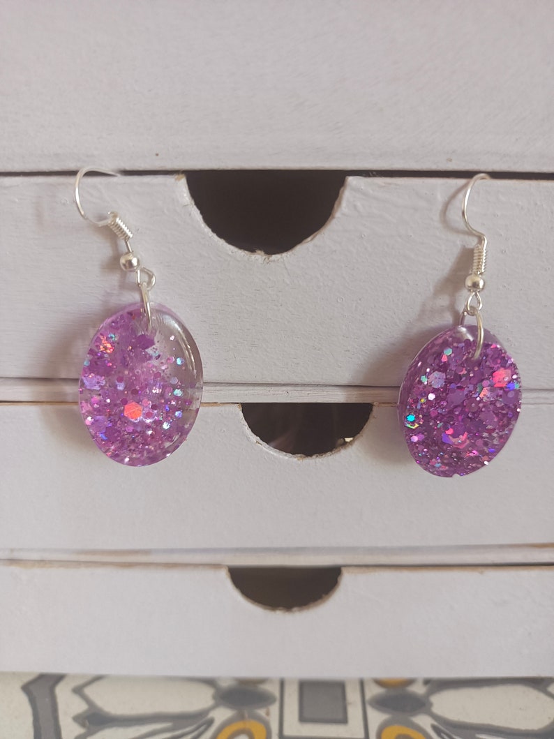 May include: A pair of silver dangle earrings with oval-shaped purple resin charms filled with iridescent glitter.