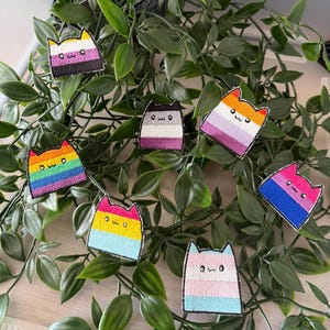 Könnte beinhalten: Eine Sammlung von bestickten Katzen-Patches mit verschiedenen LGBTQ+-Pride-Flaggen-Designs. Jeder Patch zeigt eine Katzenkontur mit dem Ausdruck "owo" und einem anderen Flaggenmuster, darunter Regenbogen-, pansexuelle und Transgender-Flaggen. Die Patches sind vor einem Hintergrund aus grünem Laub angeordnet.