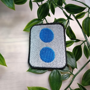 Könnte beinhalten: Ein weißer und grauer gestickter Aufnäher mit zwei blauen Kreisen. Der Aufnäher hat einen schwarzen Rand.