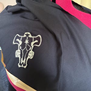 Op de afbeelding: Zwarte stof met een capuchon en een goud en zwart embleem. Het embleem is een gestileerde stierenkop met een ster in het midden. De stof is gedrapeerd over een rode paspop.