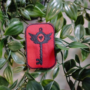 May include: A red embroidered patch featuring a black key design. The key has wings and a heart at its centre. The patch is bordered in black.