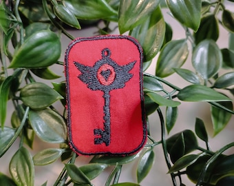May include: A red embroidered patch with a black key design. The key has wings and a heart in the center. The patch is surrounded by a black border.