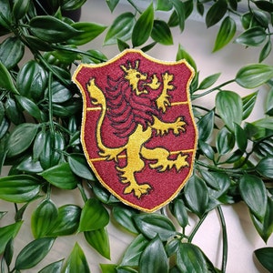May include: A red and gold embroidered patch depicting a lion with a crown on its head. The lion is standing on its hind legs with its claws extended. The patch is shaped like a shield and has a red background with gold trim.