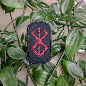 May include: A black embroidered patch with a red iron cross symbol. The patch is surrounded by a black border.