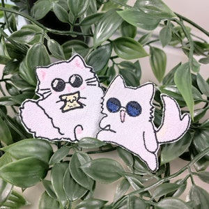 Könnte beinhalten: Zwei weiße, gestickte Bügelbilder von Comic-Katzen mit Sonnenbrillen. Eine Katze hält ein Stück Essen.